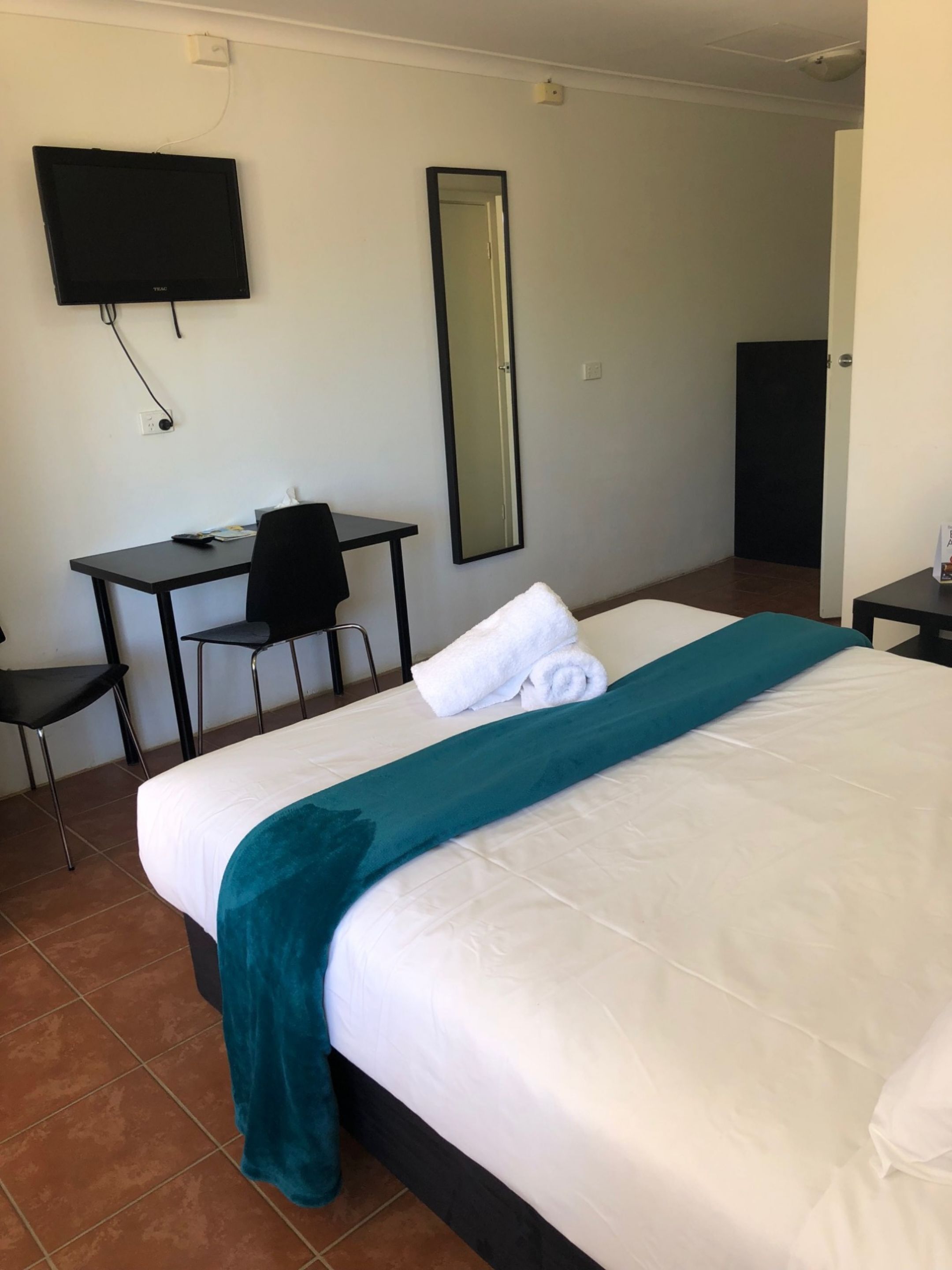 A hotel room with a bed, a desk, a chair, a TV, and a mirror.