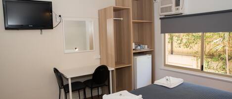 A hotel room with a bed, a desk with a chair, a television, a wardrobe, a mini-fridge, and a window with a view of greenery.