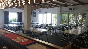 Un vaste espace dédié à la salle à manger, avec de nombreuses tables et chaises, de grandes fenêtres laissant entrer la lumière naturelle et des luminaires au plafond.