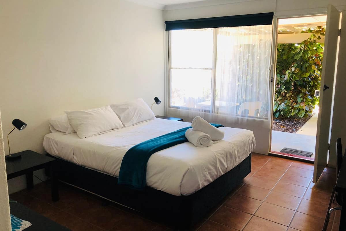 Un dormitorio con cama, mesita de noche, ventana con cortinas y vistas a la vegetación.