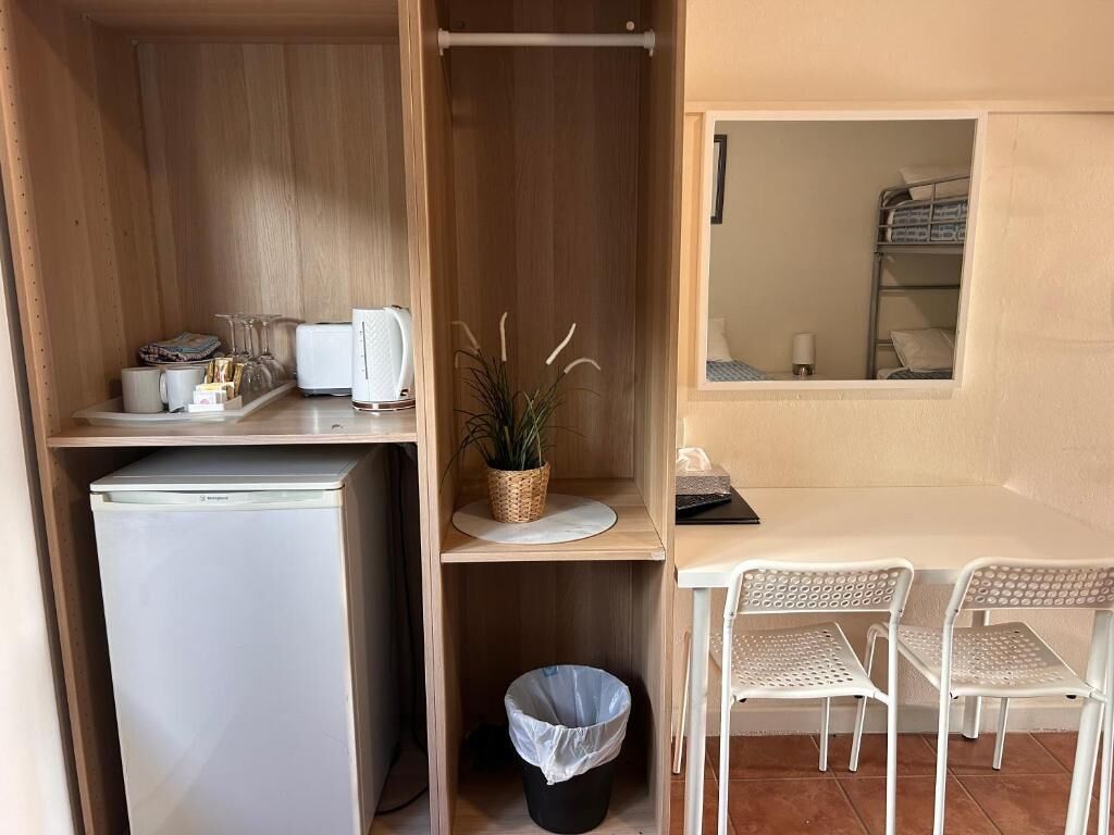 A compact room with a kitchenette, a small trash bin, a dining area with a table and chairs, and a window with a view of a bunk bed.