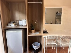 Una habitación compacta con kitchenette, un cesto de basura pequeño, una zona de comedor con mesa y sillas, y una ventana con vista a una cama individual.