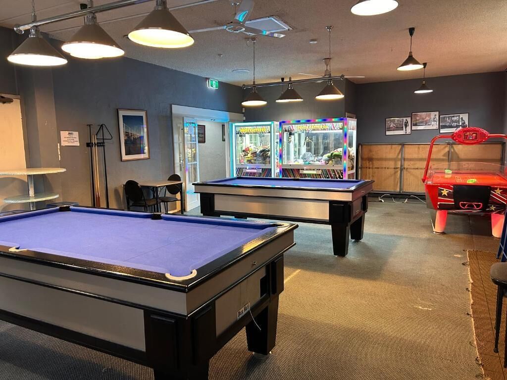 A pool table, two arcade machines, and a red arcade cabinet in a room with ceiling fans and framed pictures on the wall.