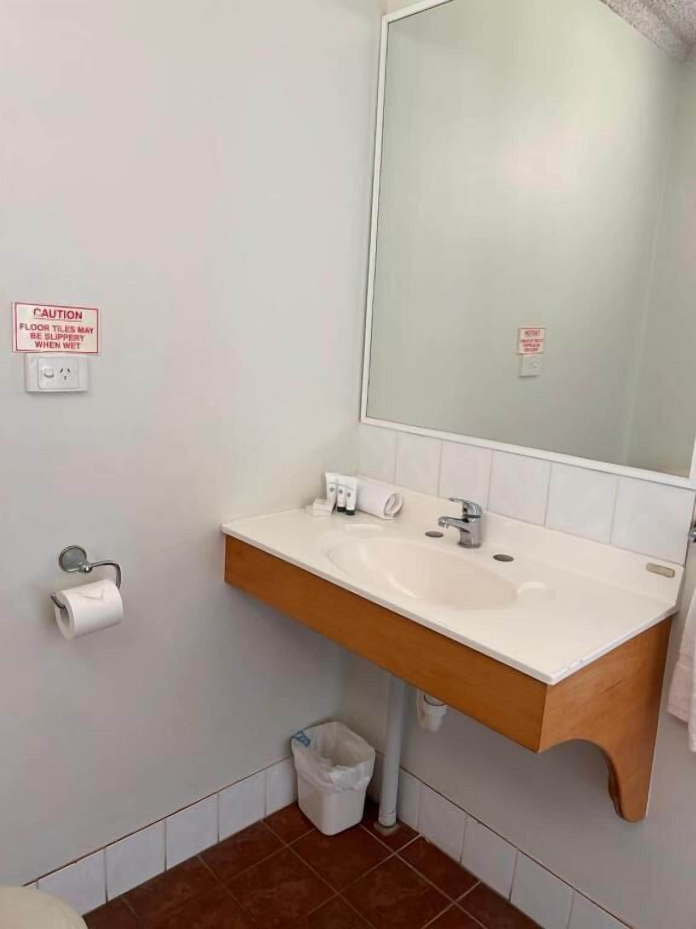 A bathroom with a white sink, wooden vanity, and a mirror. There are caution signs on the wall and a trash bin below the sink.