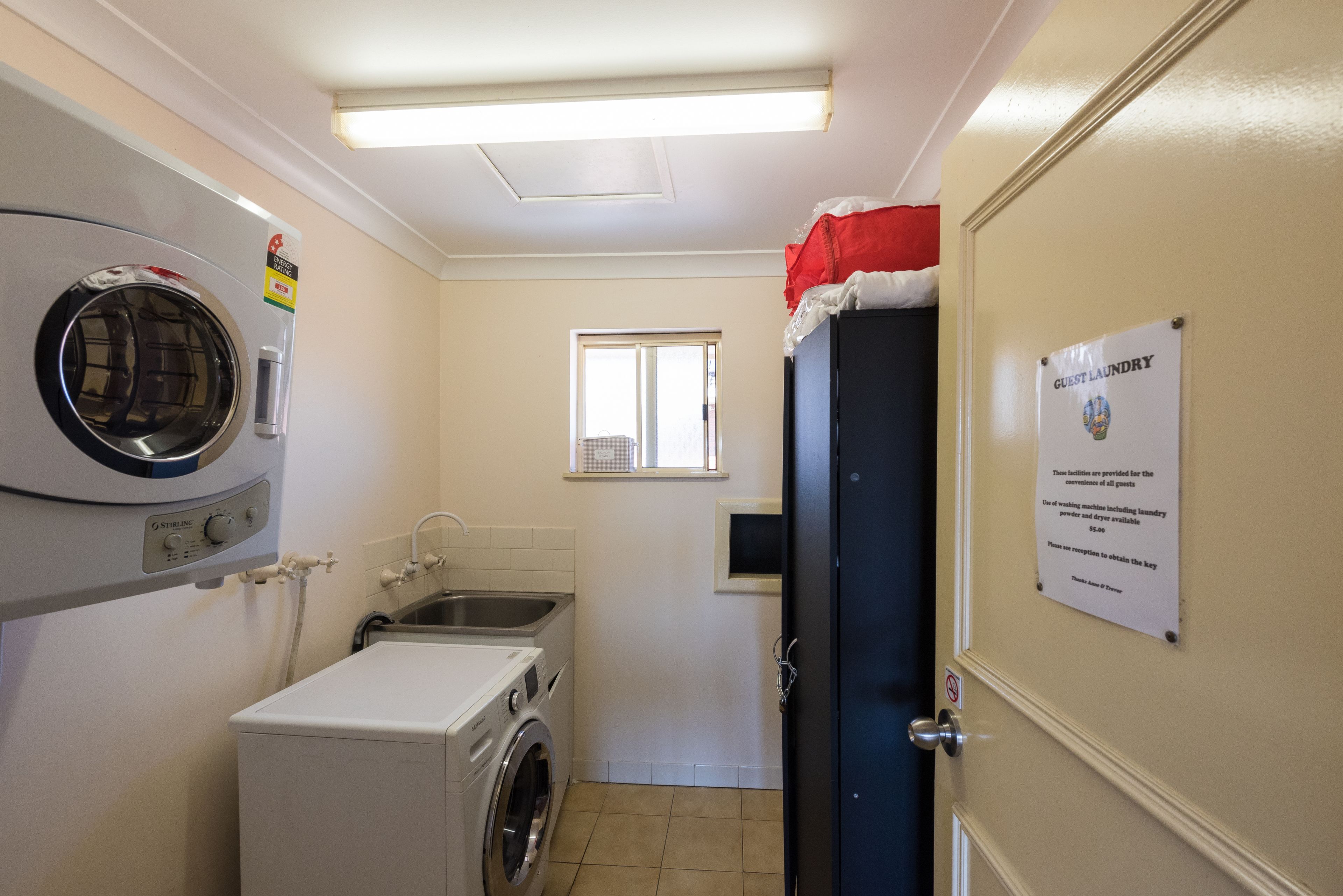 Zona de lavandería con lavadora, secadora y lavamanos. Hay una ventana y una puerta con un letrero que indica servicios de lavandería para huéspedes.