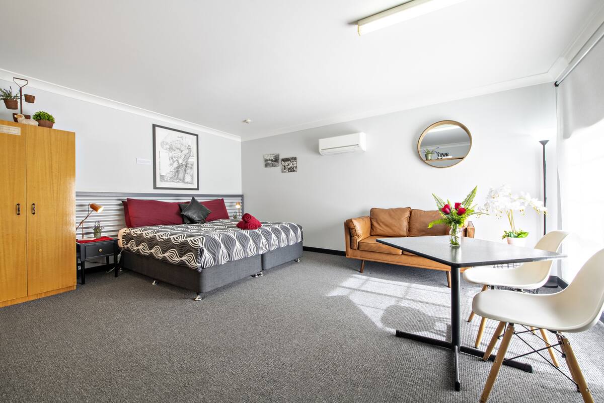A modern hotel room with a bed, a sofa, a dining table with chairs, a wooden wardrobe, and a small desk with a lamp.