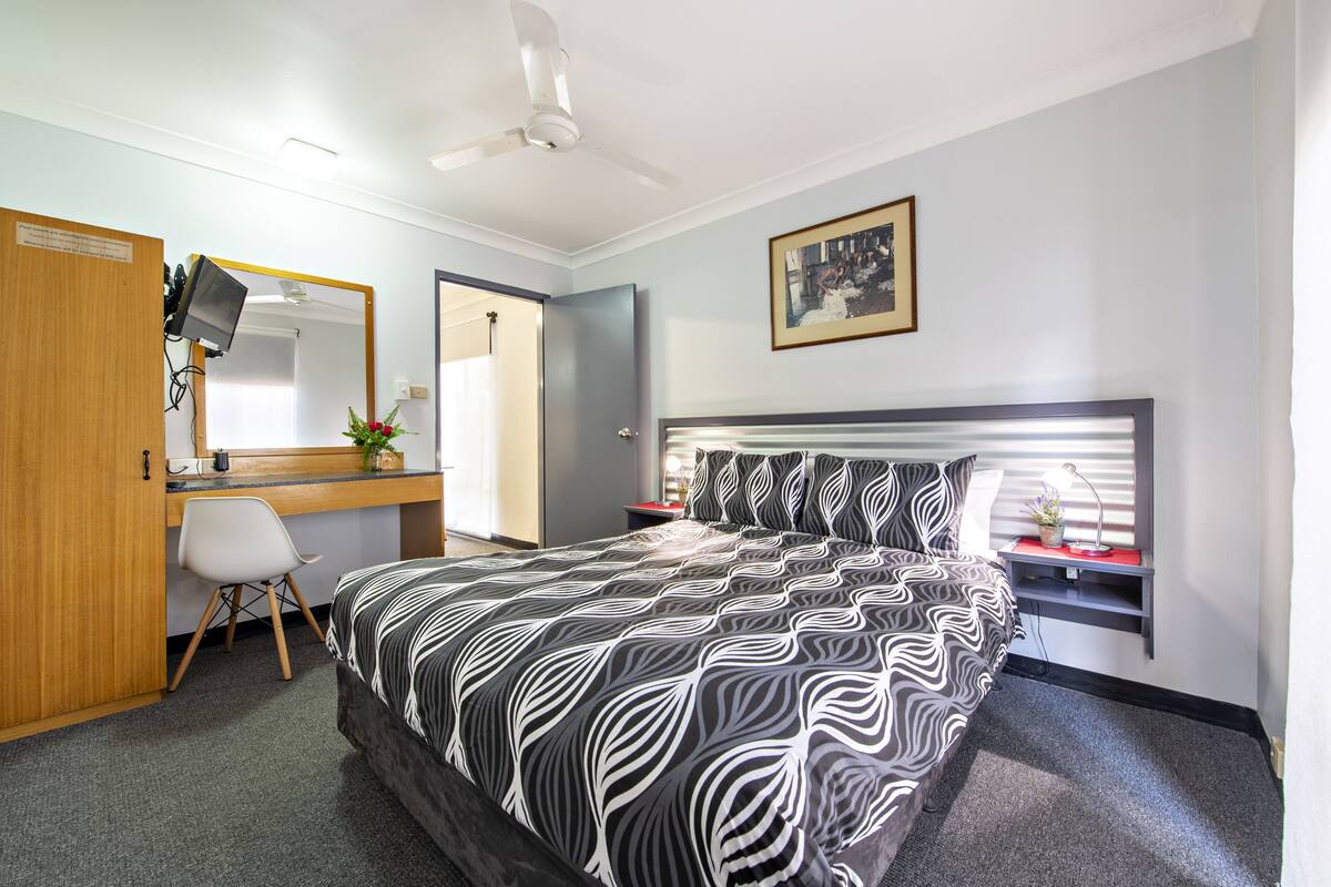 A hotel room with a bed, a desk, a chair, a mirror, a ceiling fan, and a picture on the wall.