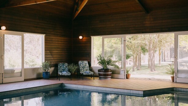 An indoor swimming pool with wooden beams, potted plants, and two doors leading to a garden. - Flowerdale Estate (Strath Creek)
