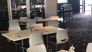 A modern dining area with white tables and chairs, a carpeted floor, and a view of the outside through large windows. - The Premier Hotel (Broadmeadow)