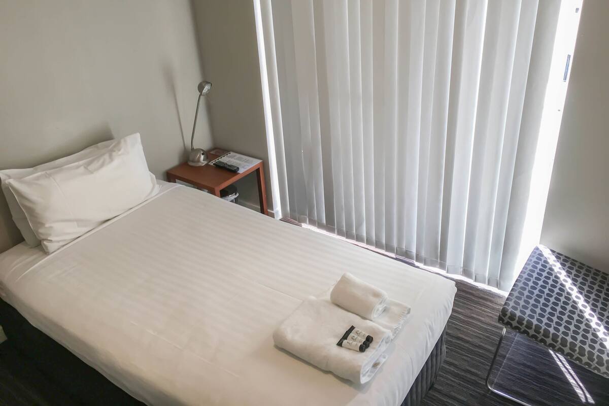 A hotel room with a neatly made bed, a bedside table with a lamp, and a window with blinds.