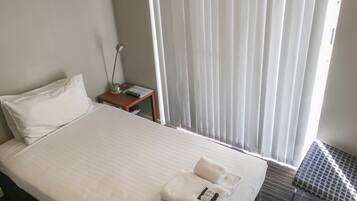 A hotel room with a neatly made bed, a bedside table with a lamp, and a window with blinds.