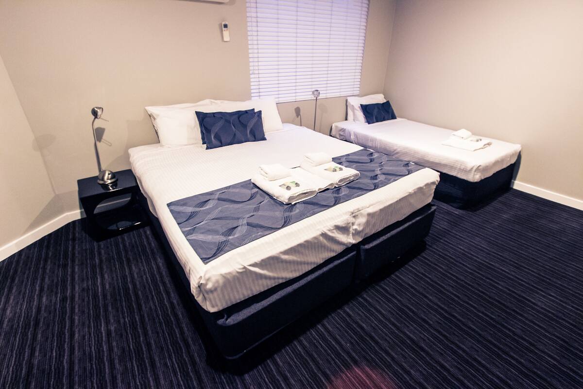 A hotel room with two beds, a nightstand with a lamp, and a window with blinds.