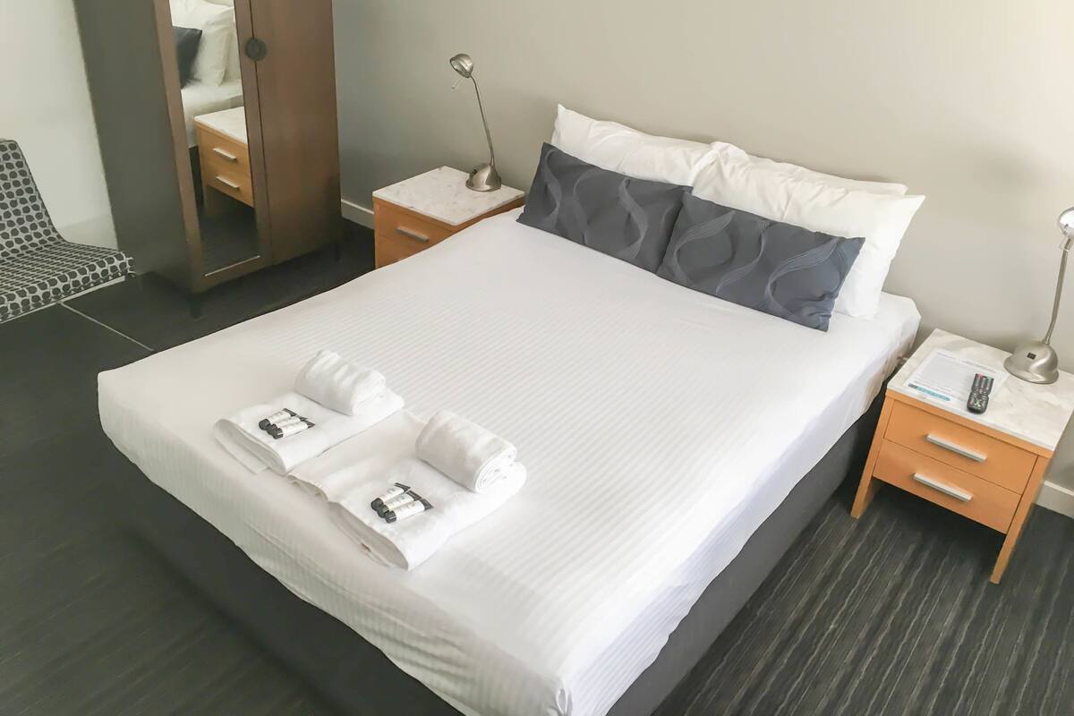 A neatly made bed with white linens and two towels, a bedside table with a lamp, a nightstand with a remote control, and a mirror on the wall.