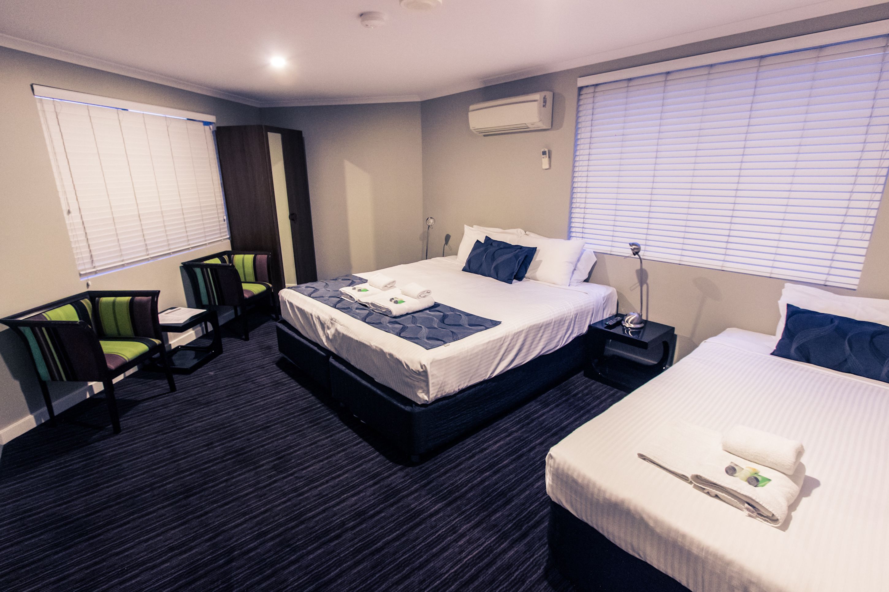 Una habitación de hotel con una cama grande, una cama más pequeña, un escritorio con silla y una ventana con persianas.