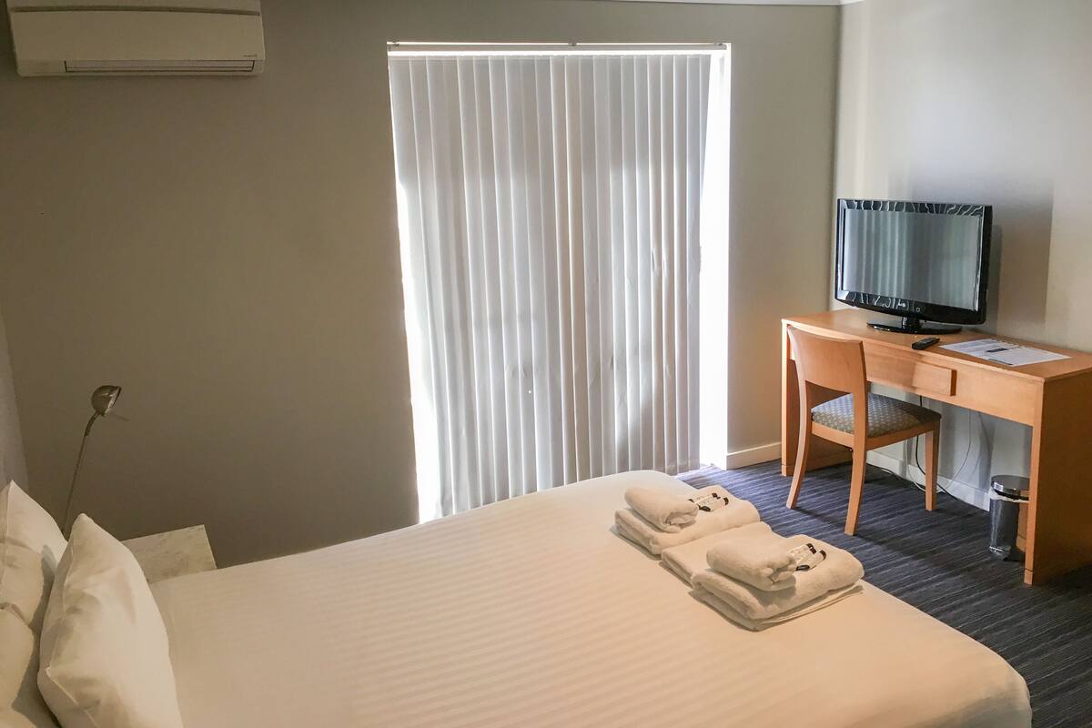 A hotel room with a bed, a desk with a television, a chair, and a window with blinds.
