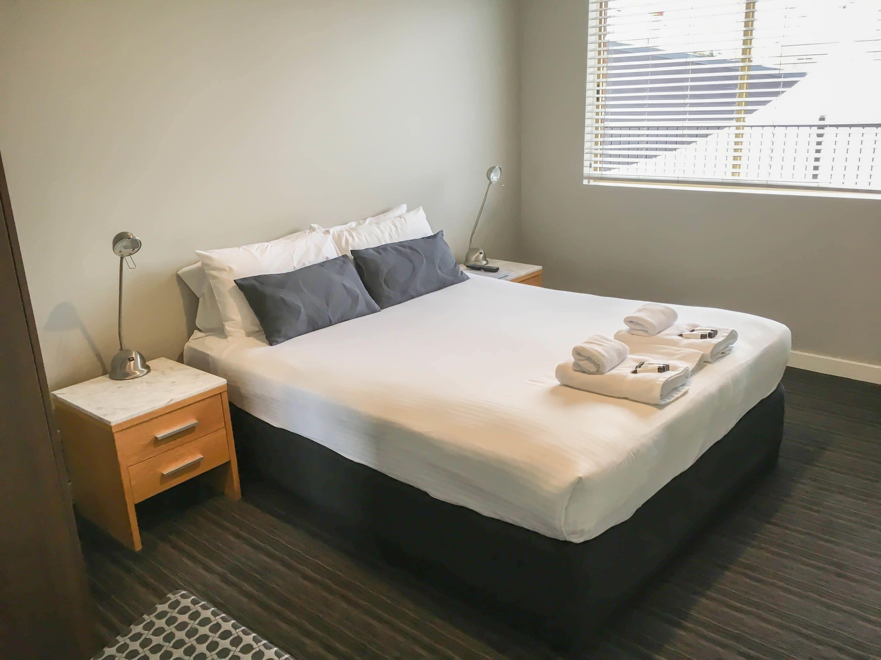 Een netjes opgemaakt bed met witte lakens en twee kussens, een nachtkastje met een lamp, en een raam met jaloezieën.