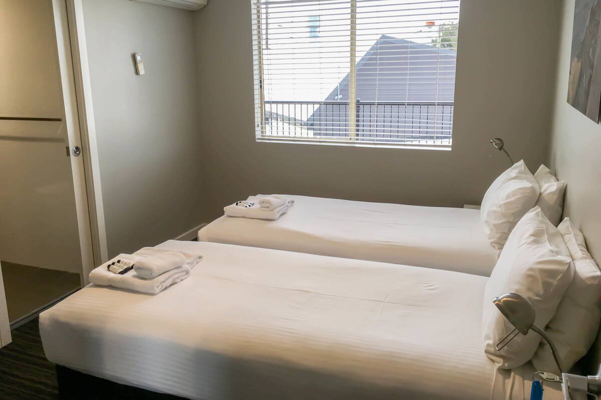 A hotel room with two beds, a window with blinds, a door, and a wall-mounted air conditioner.