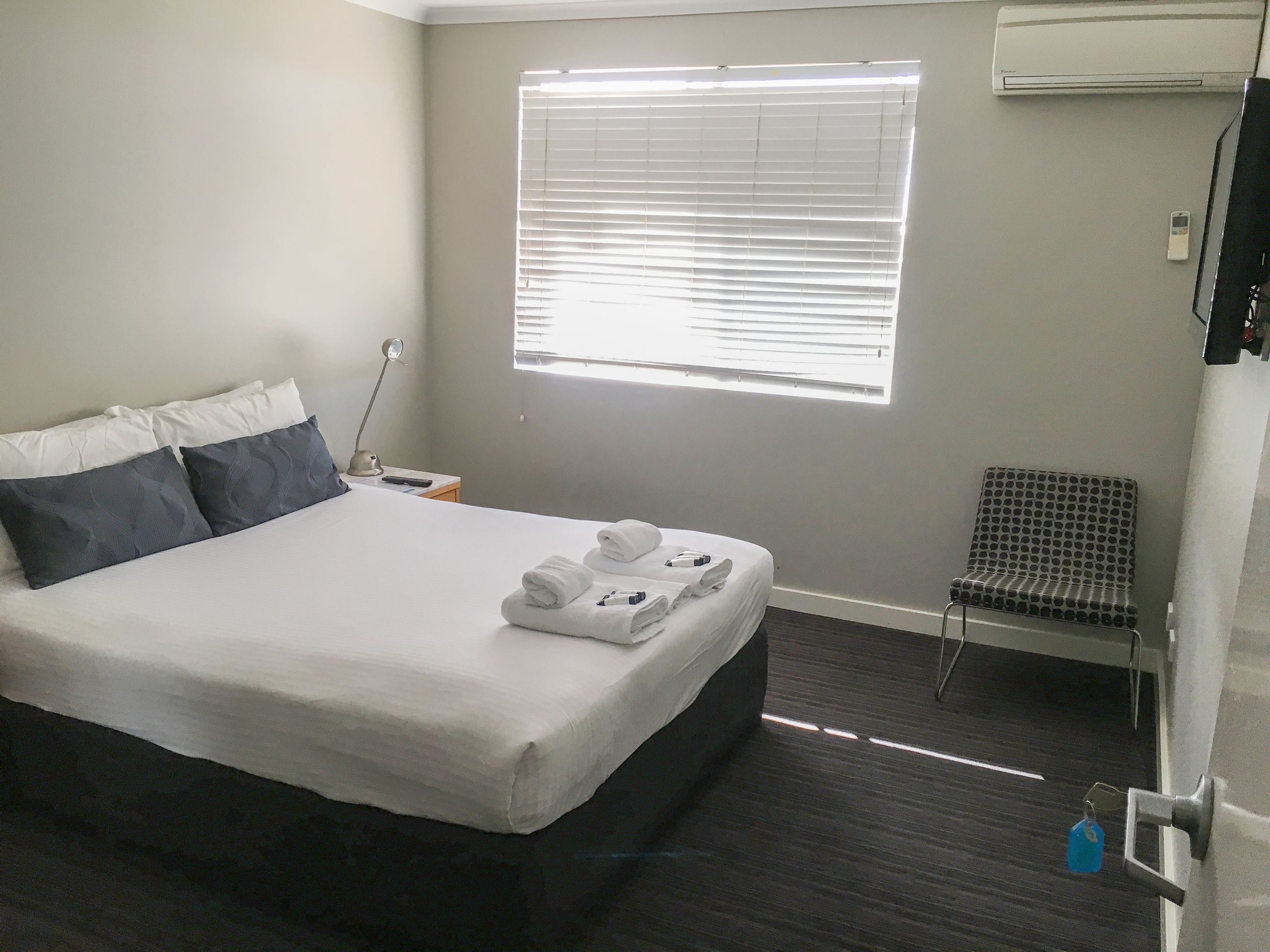 A hotel room with a bed, a chair, a desk with a lamp, and an air conditioning unit.