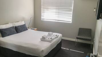 A hotel room with a bed, a chair, a desk with a lamp, and an air conditioning unit.