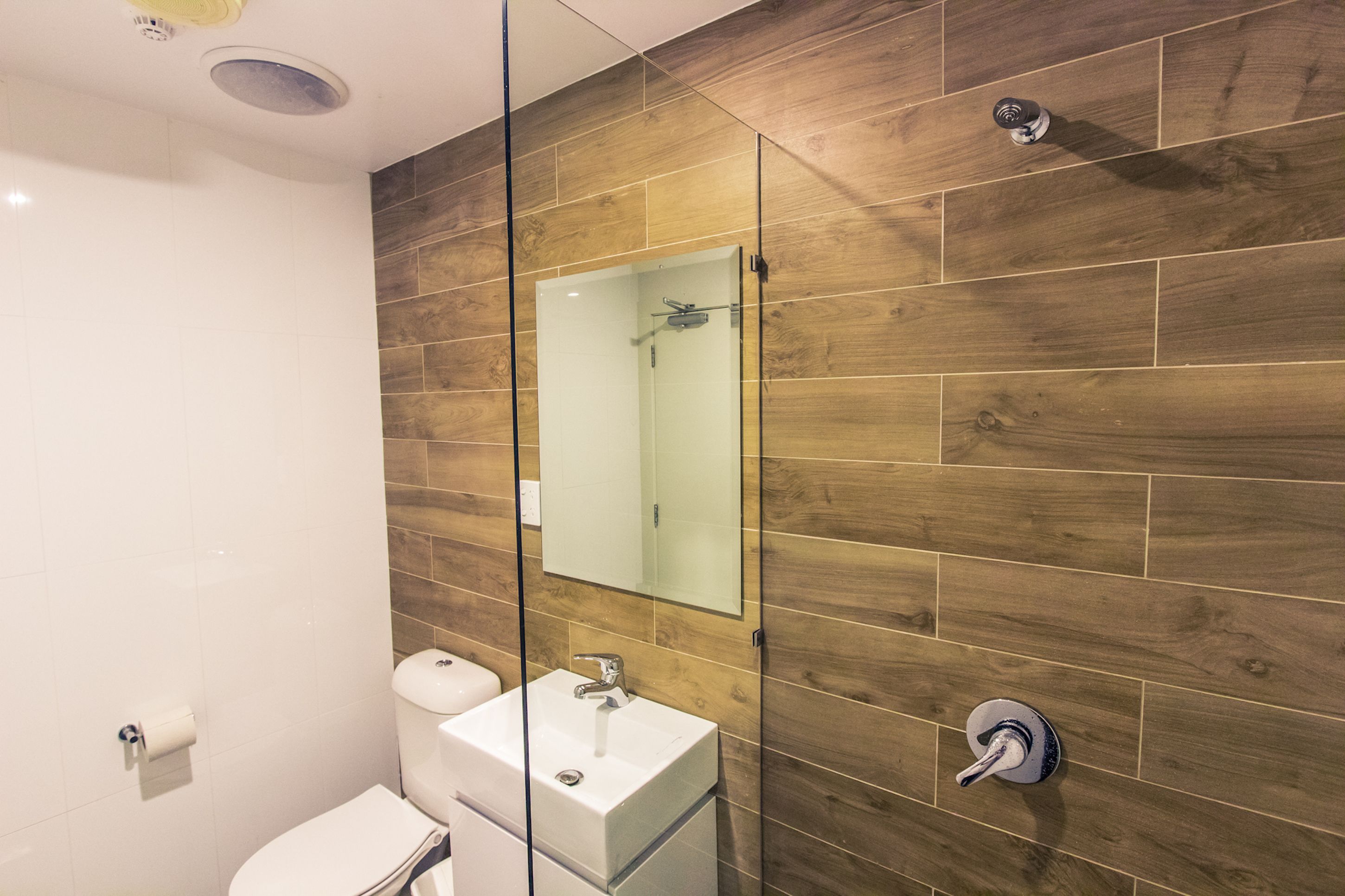 A modern bathroom with a wooden wall, a glass shower enclosure, a white sink, and a toilet.