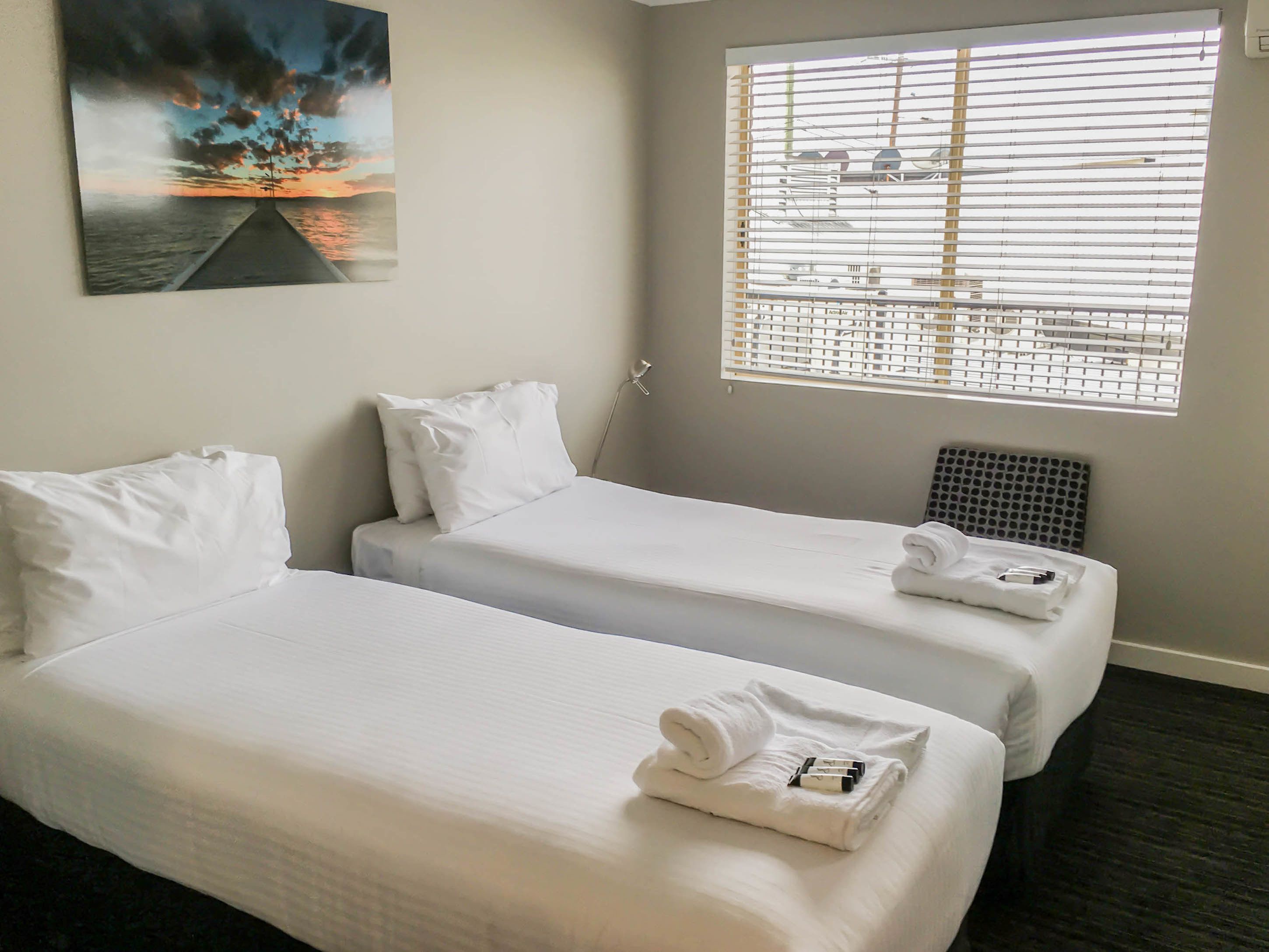 A hotel room with two single beds, white bedding, a painting of a sunset, and a window with blinds.