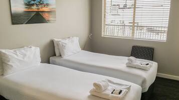 A hotel room with two single beds, white bedding, a painting of a sunset, and a window with blinds.