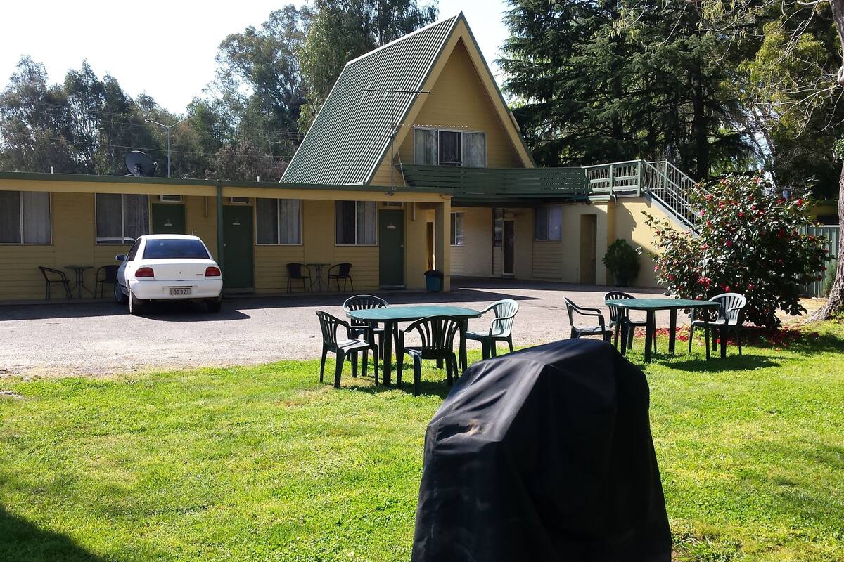 Un edificio con techo inclinado, un coche estacionado adelante, mesas al aire libre y un árbol.