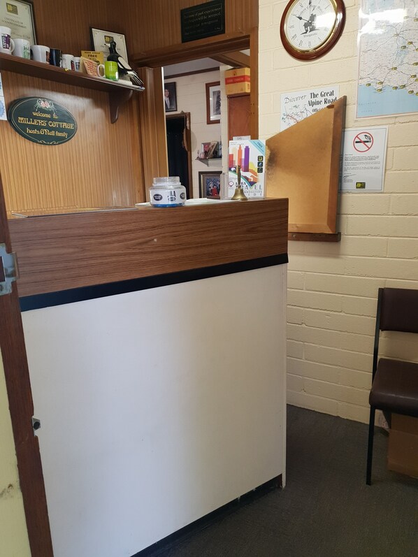 A reception desk with a wooden counter and a white panel, surrounded by various items like a clock, a framed certificate, and a map on the wall. - Millers Cottage Motel (Wangaratta)