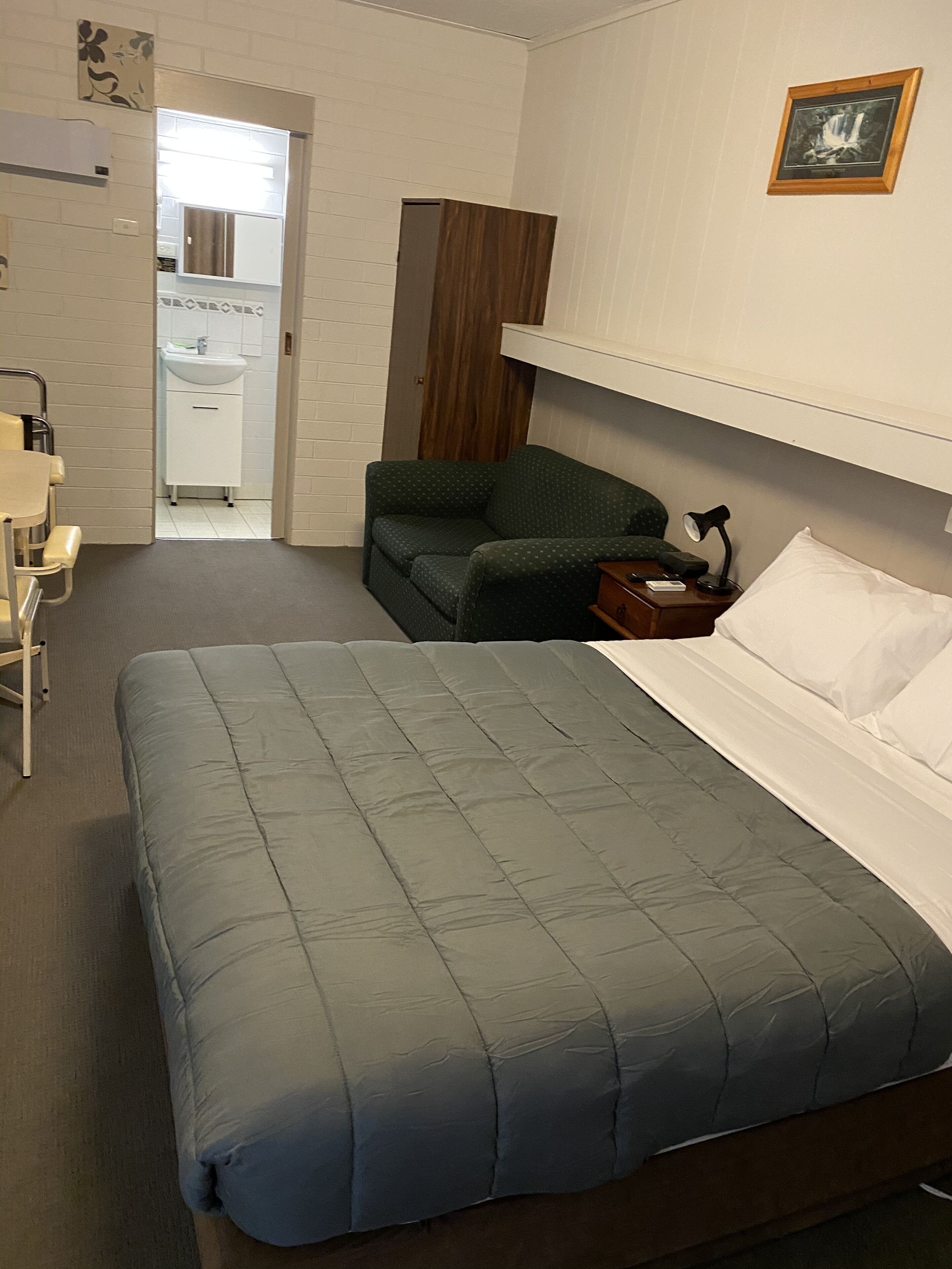 Habitación de hotel ordenada con cama, una silla, mesita de noche con lámpara y un mueble bajo.