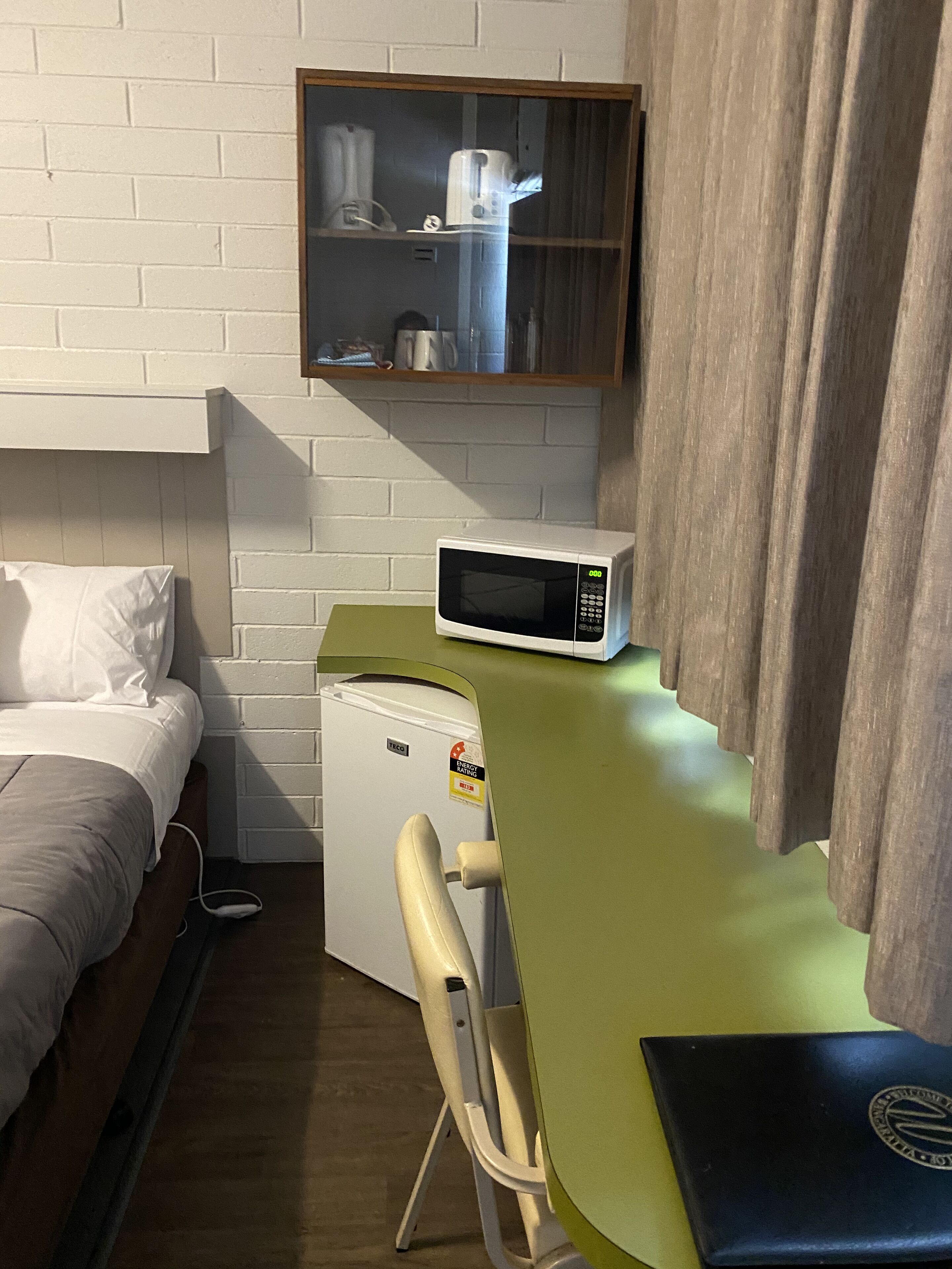 Habitación de hotel compacta con una cama, un pequeño kitchenette, un microondas, una silla y un estante con utensilios de cocina.