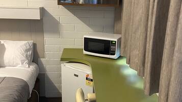 Habitación de hotel compacta con una cama, un pequeño kitchenette, un microondas, una silla y un estante con utensilios de cocina.