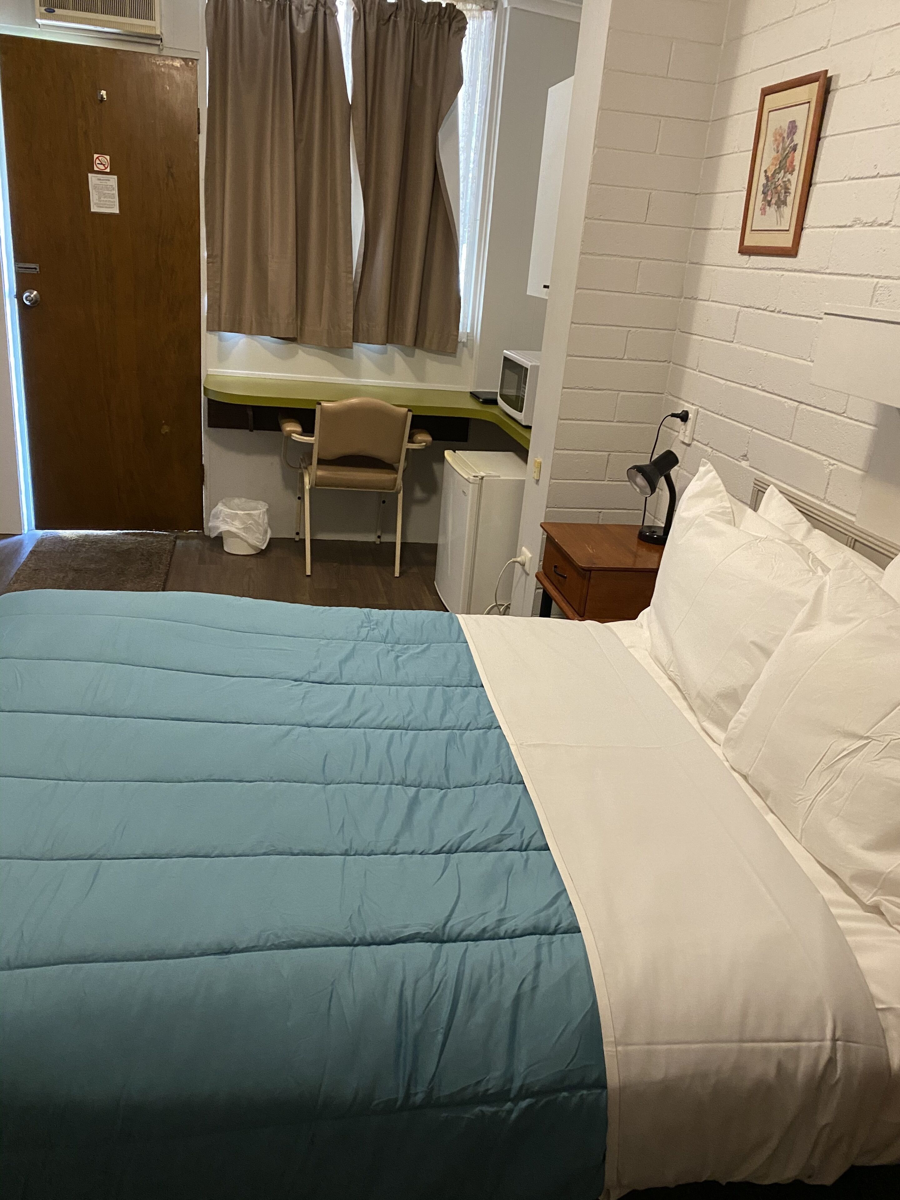 A bedroom with a bed, a desk, a chair, a small refrigerator, and a window with curtains.