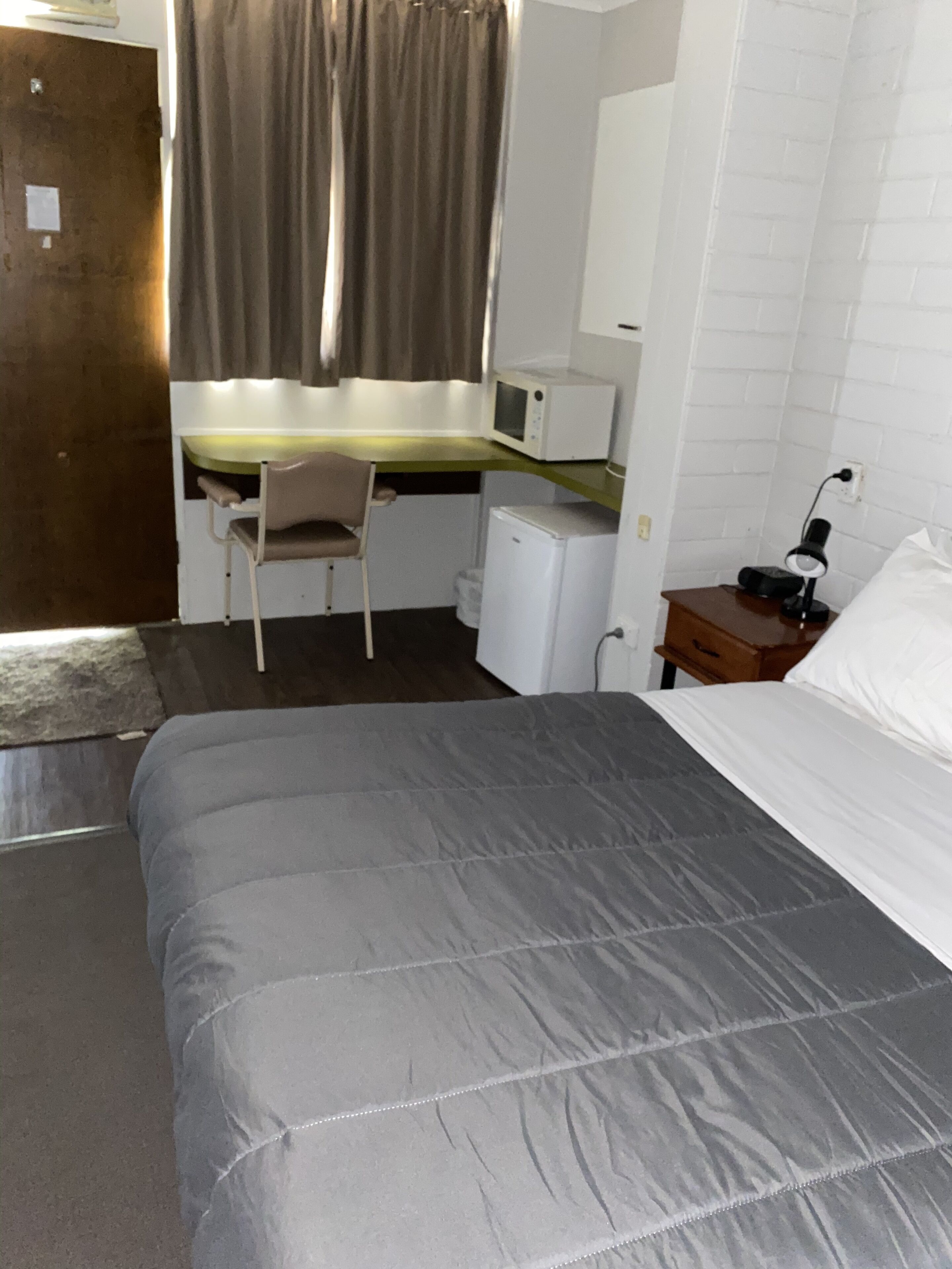 A hotel room with a bed, a desk, a chair, a microwave, a nightstand, and a lamp.