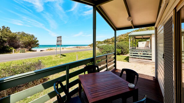 Un espace extérieur abrité, doté d’une table et de chaises en bois, qui offre une vue sur une plage et une route.