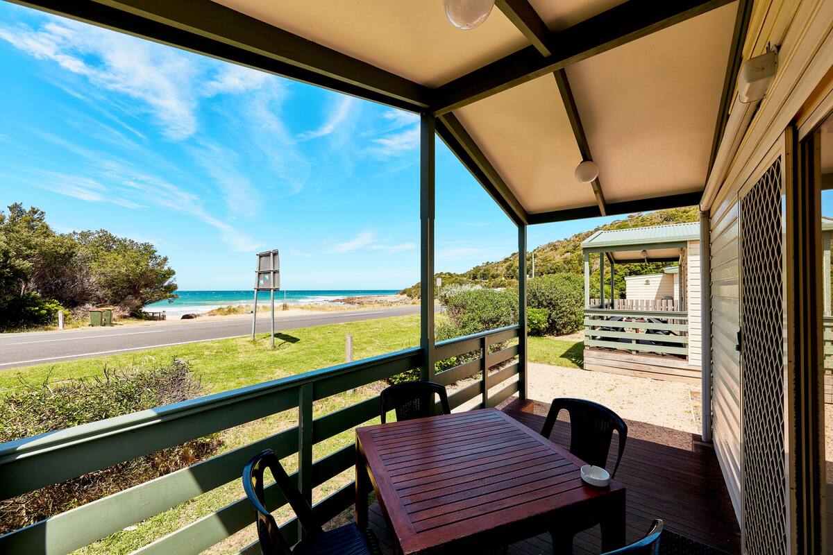 Un espacio al aire libre cubierto con una mesa y sillas de madera, con vistas a una playa y una carretera.