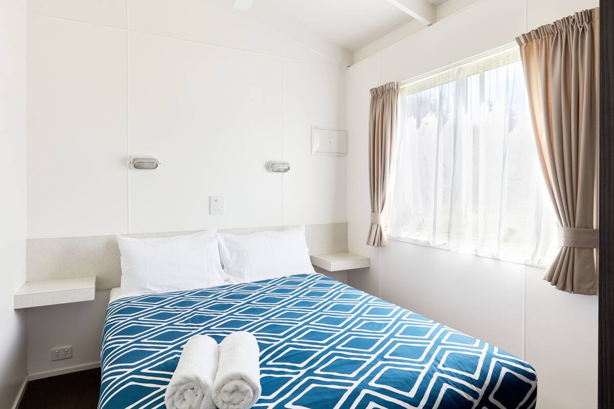 Habitación de hotel con cama, mesita de noche, ventana con cortinas y una lámpara fijada a la pared.