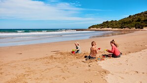 Une plage où trois enfants jouent dans le sable, un ciel dégagé et un paysage vallonné en arrière-plan.
