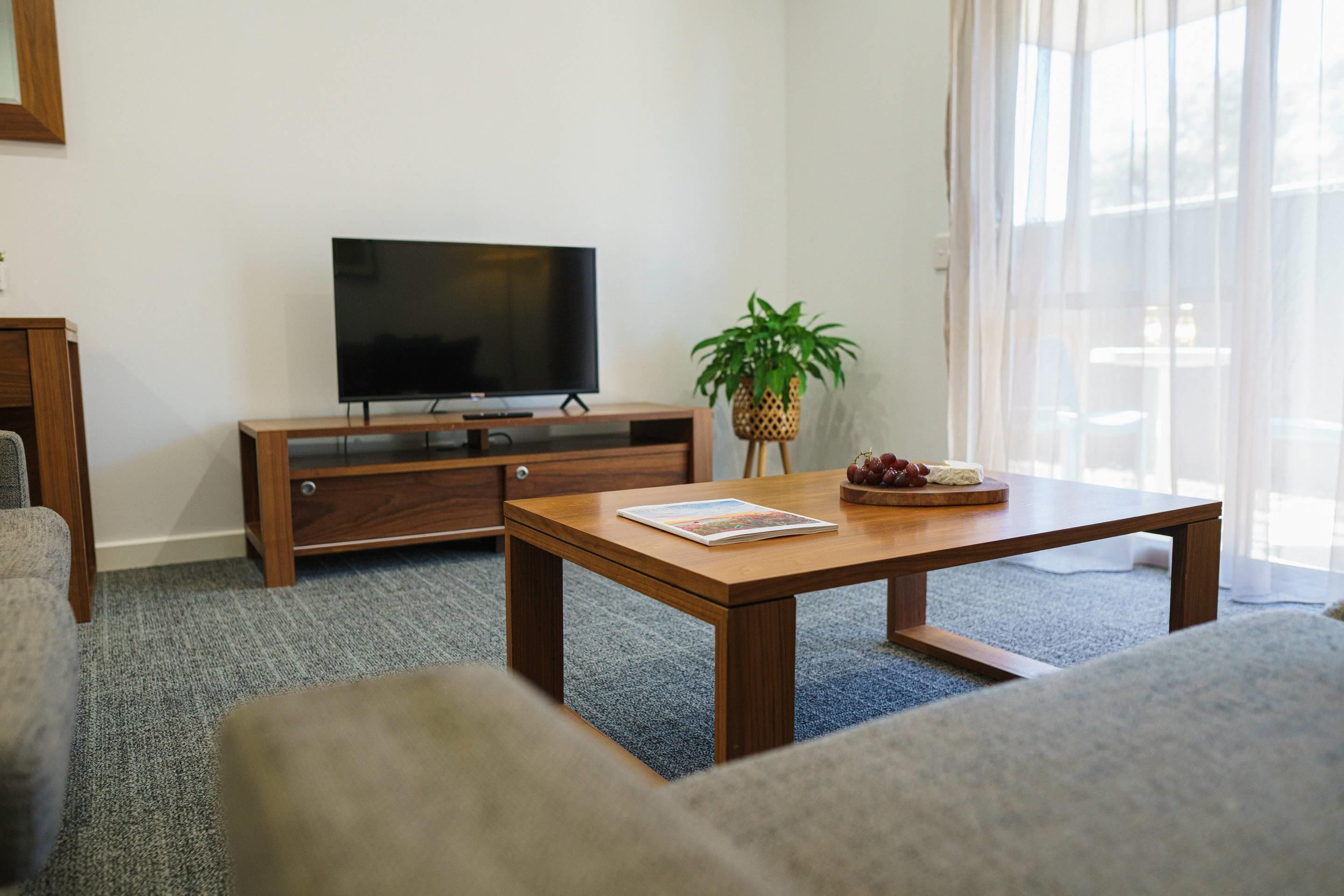 Una sala de estar con un mueble bajo para televisión de madera, un televisor de pantalla plana, una mesa de centro de madera con una revista, una planta y una bandeja de frutas.