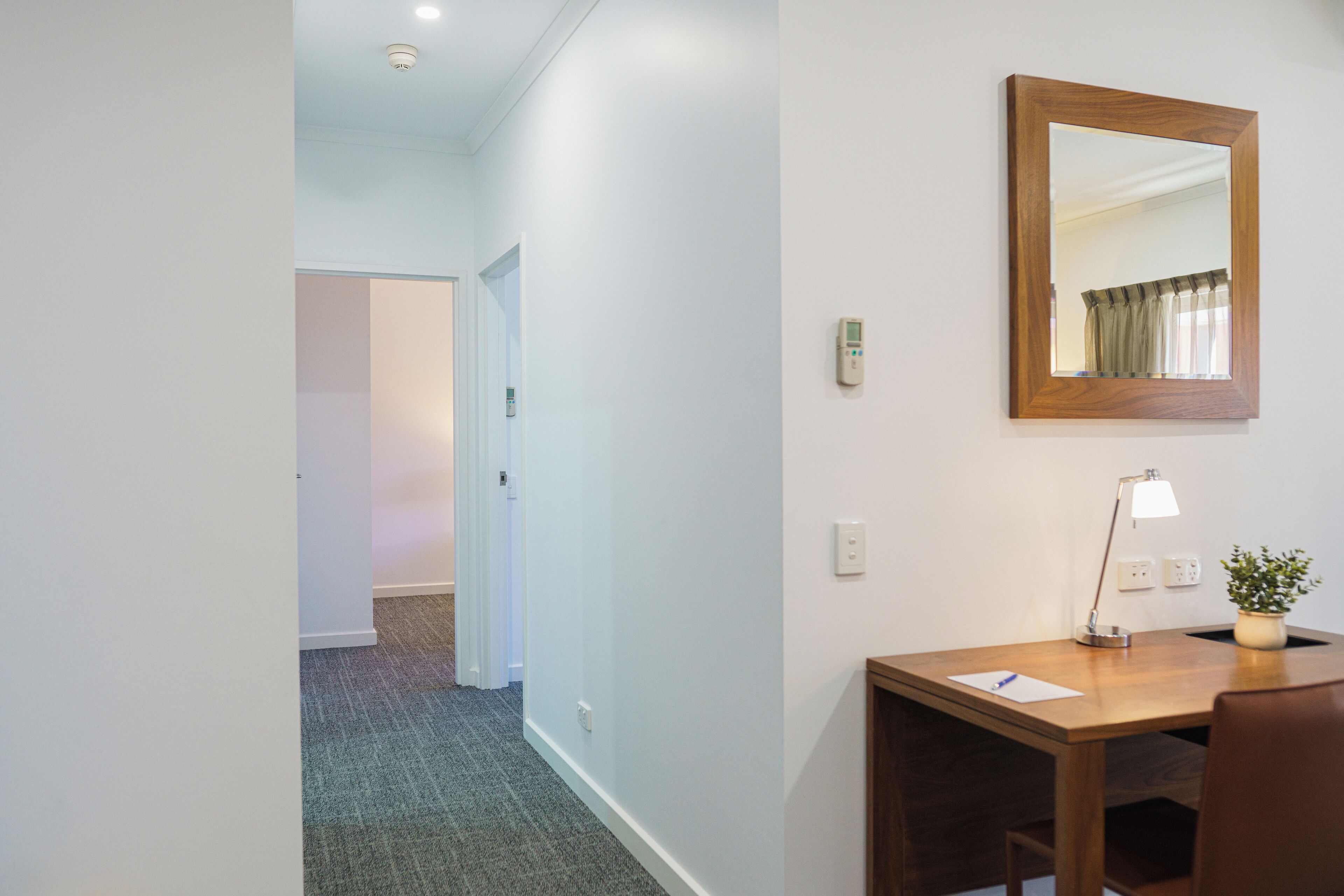 Une chambre d’hôtel avec un bureau en bois, un miroir, une lampe et une plante en pot.