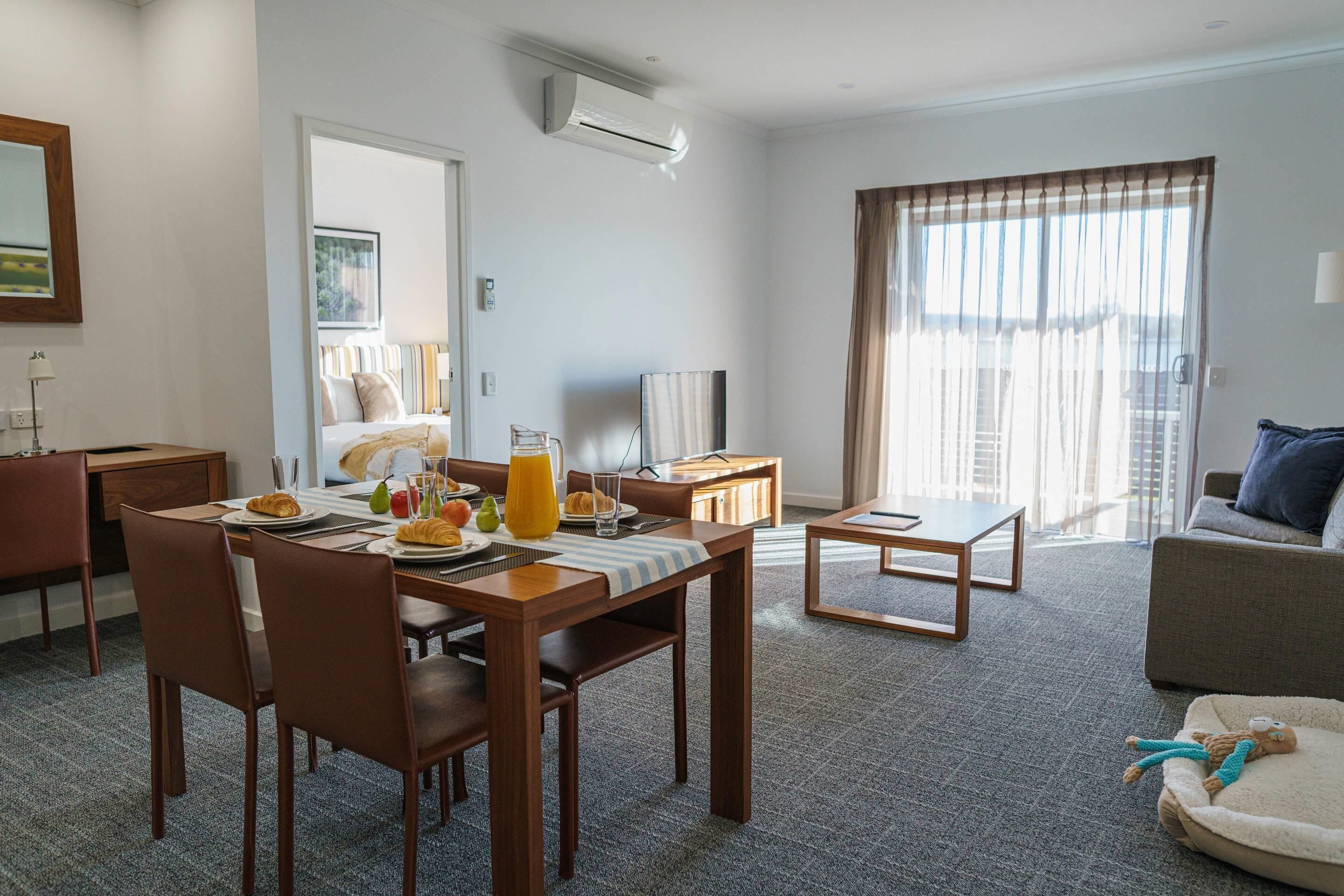 Habitación de hotel con comedor, sofá, televisor y un amplio ventanal con cortinas.