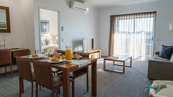 Habitación de hotel con comedor, sofá, televisor y un amplio ventanal con cortinas.