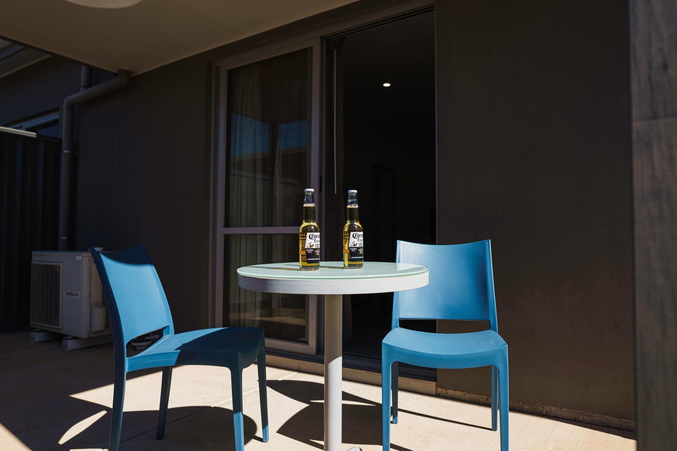 A round table with two blue chairs, two bottles of Corona beer, and an air conditioning unit in the background.