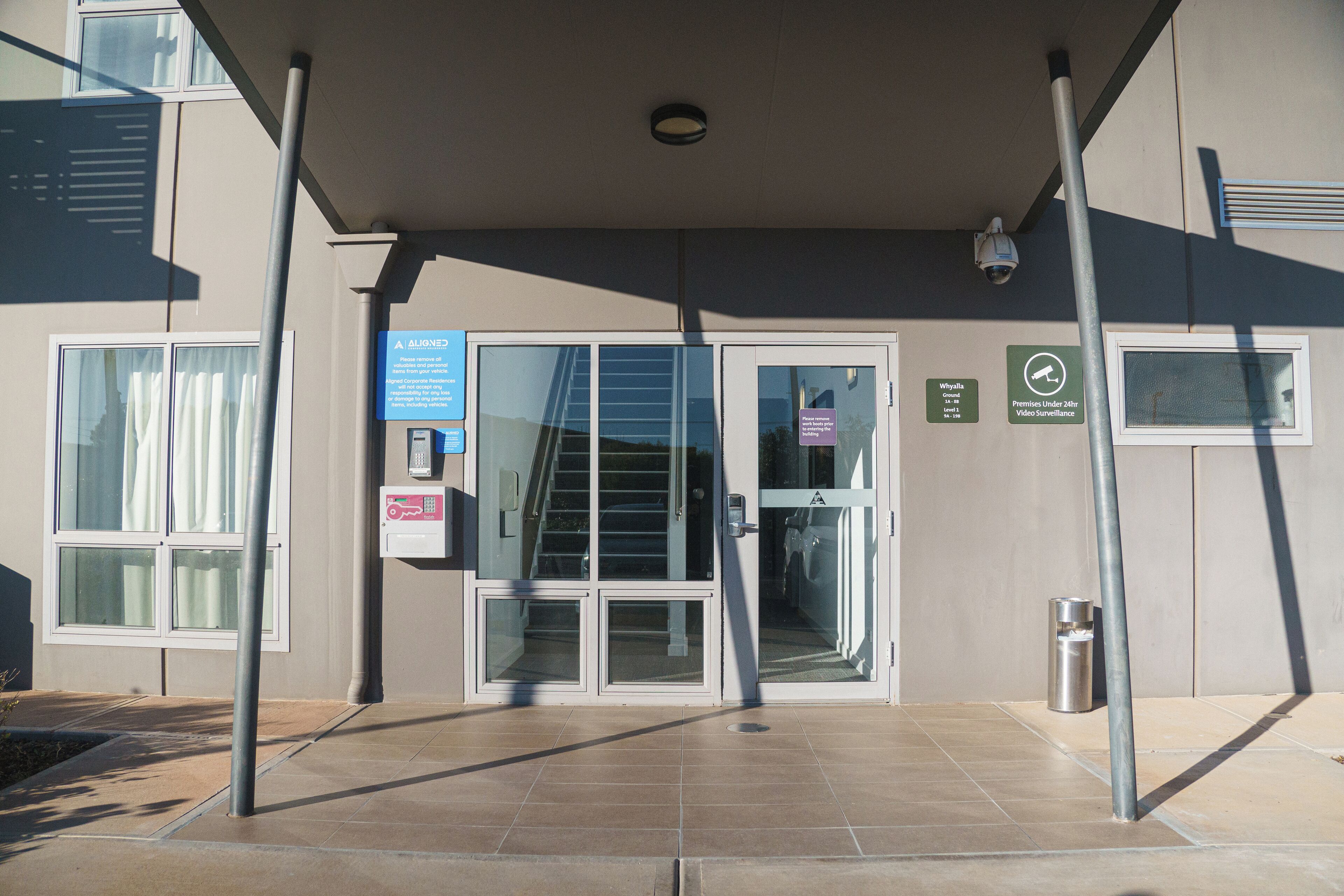 A building entrance with a covered walkway, glass doors, and a trash bin.