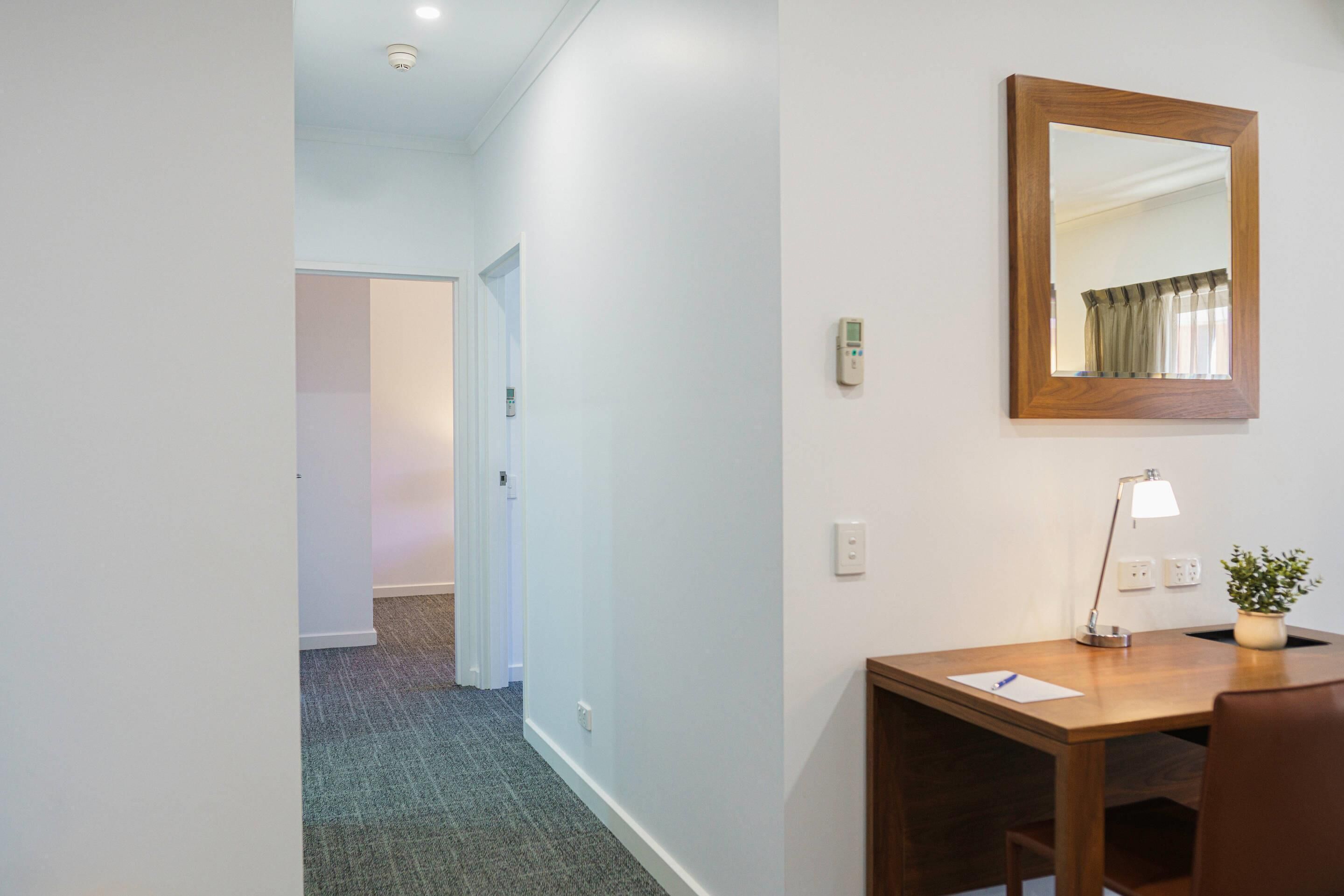 A hotel room with a desk, chair, lamp, and a mirror on the wall.