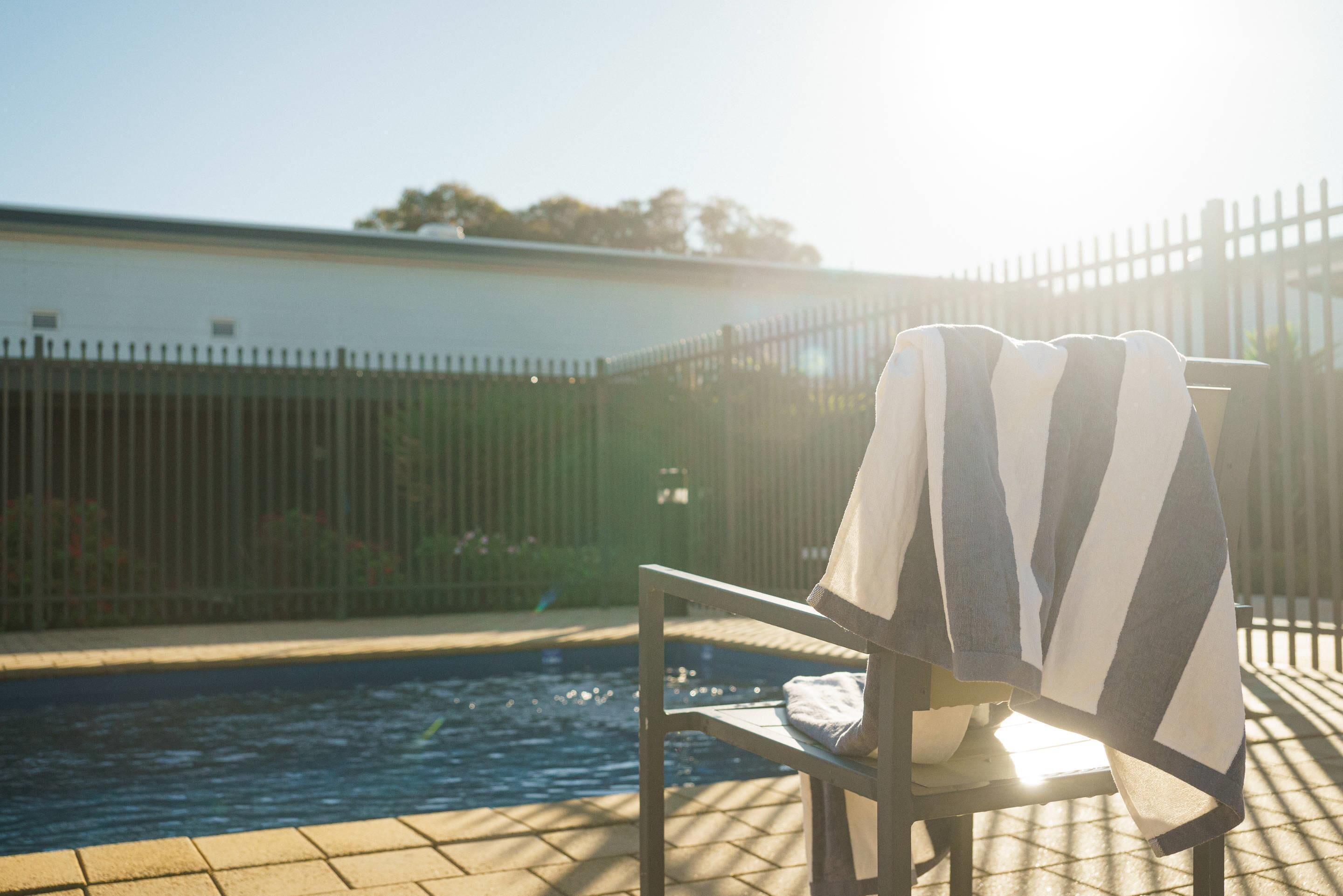 A poolside chair with a towel, a pool, and a fence in the background.