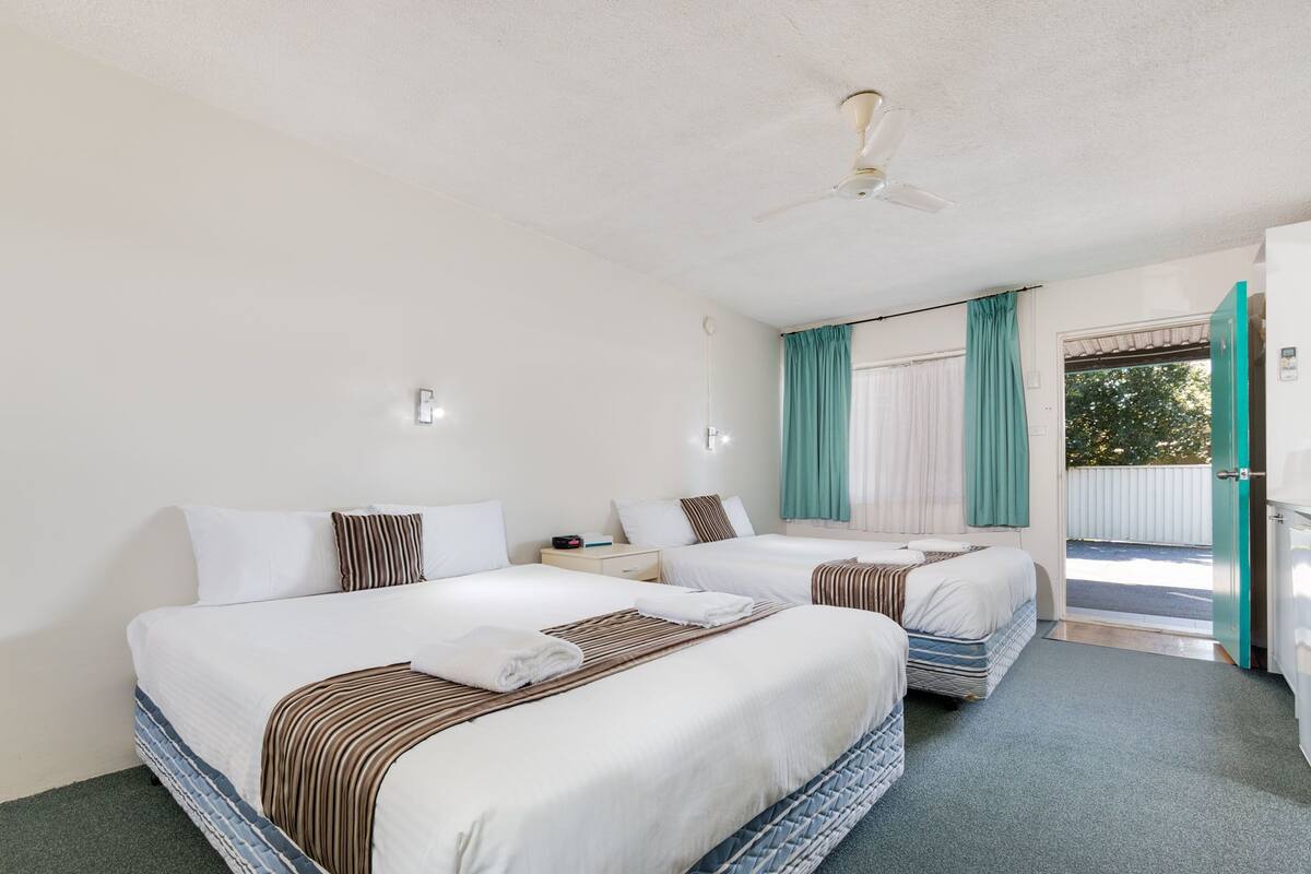 Habitación de hotel con dos camas, ventilador de techo, ventana con cortinas y puerta hacia el exterior.