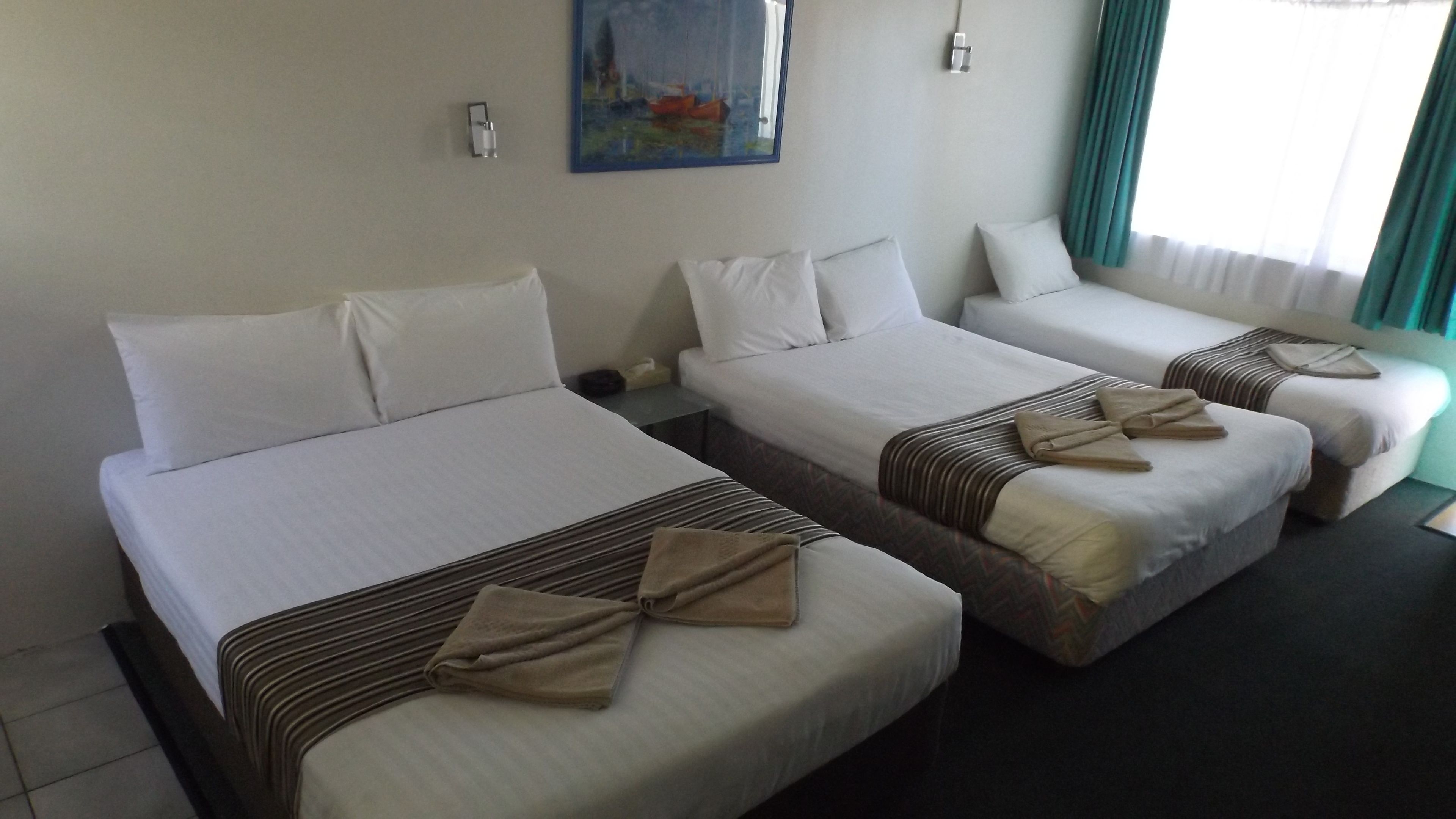 A hotel room with three single beds, each with white linens and brown decorative pillows. There is a painting on the wall and a window with curtains.