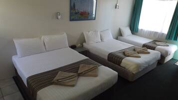 A hotel room with three single beds, each with white linens and brown decorative pillows. There is a painting on the wall and a window with curtains.