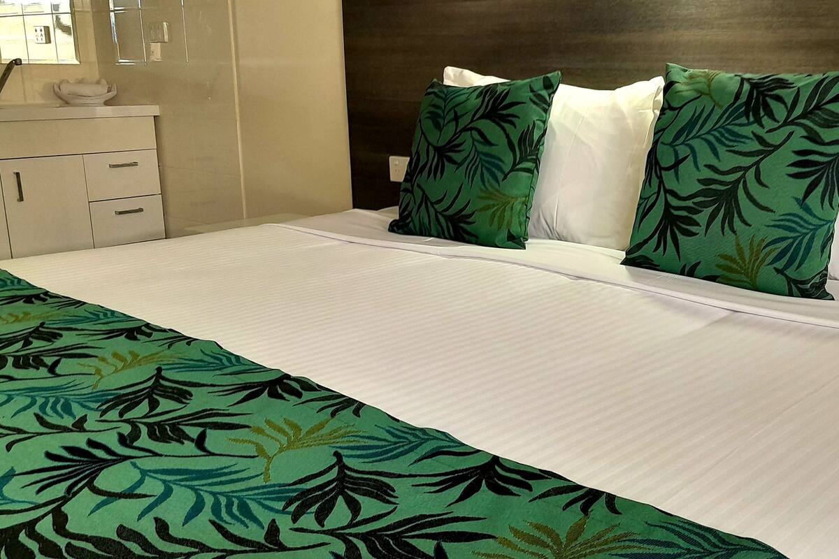 Habitación de hotel con cama, ropa de cama blanca, colcha de diseño con hojas verdes y cabecera de madera.