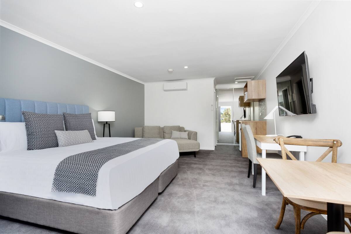 A modern hotel room with a large bed, a sofa, a desk, and a TV mounted on the wall.
